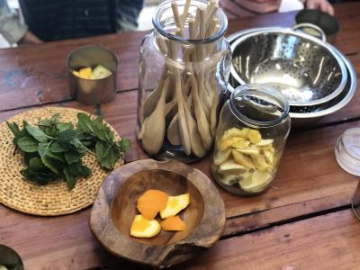 Natural ingredients including mint leaves and orange peels in bowls and jars, next to a bowl of wooden spoons and metal colander on a wooden table