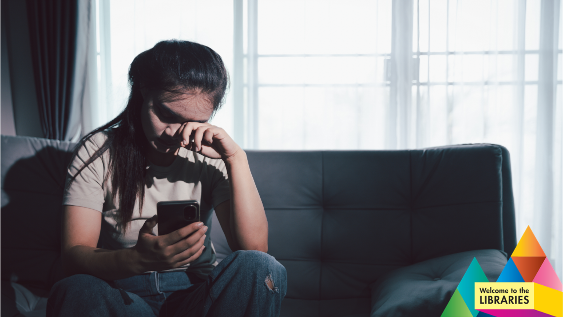 A female child/teen sits on a couch with their mobile phone in one hand, wiping their eyes with the other
