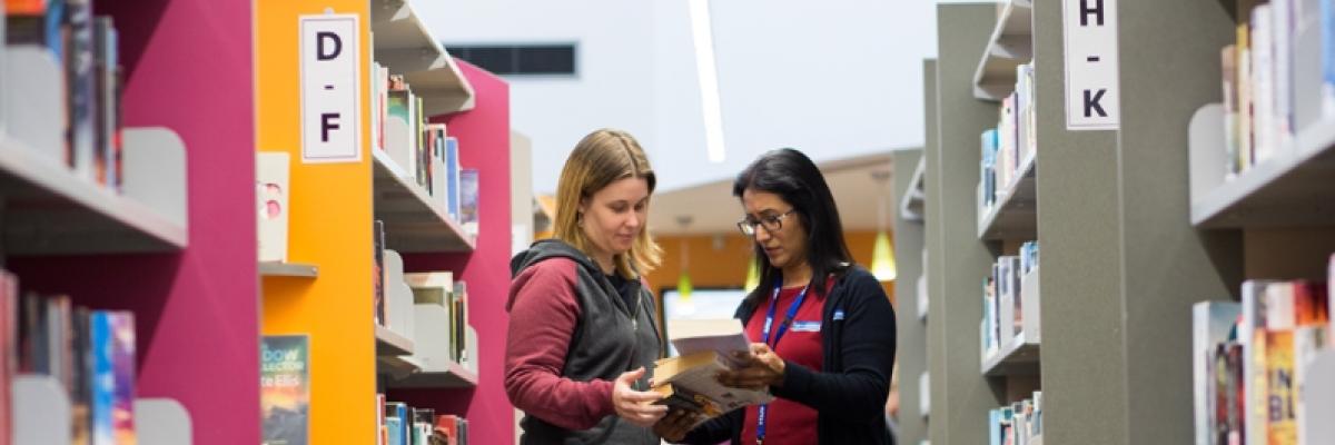 two women looking at a book in between library shelves