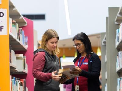 two women looking at a book in between library shelves