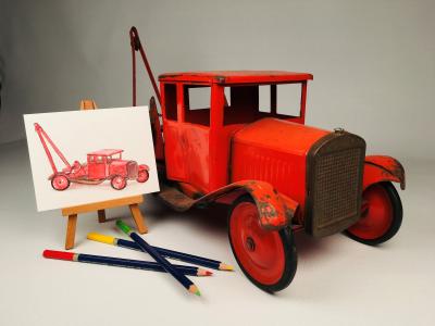 Photograph of a red toy tow truck next to an easel with a small watercolour pencil painting of the same truck.