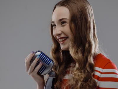 young female wearing an orange shirt with long curly hair holding a vintage style microphone in front of a grey wall.