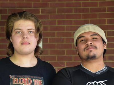 2 young males standing in front of a brick wall, one is wearing a band t-shirt and they other has a black t-shirt on with cream coloured beanie.