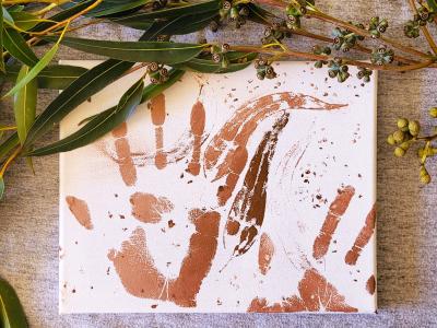 A sheet of white paper with hand prints and leaf prints in brown paint, surrounded by native Australian flora.